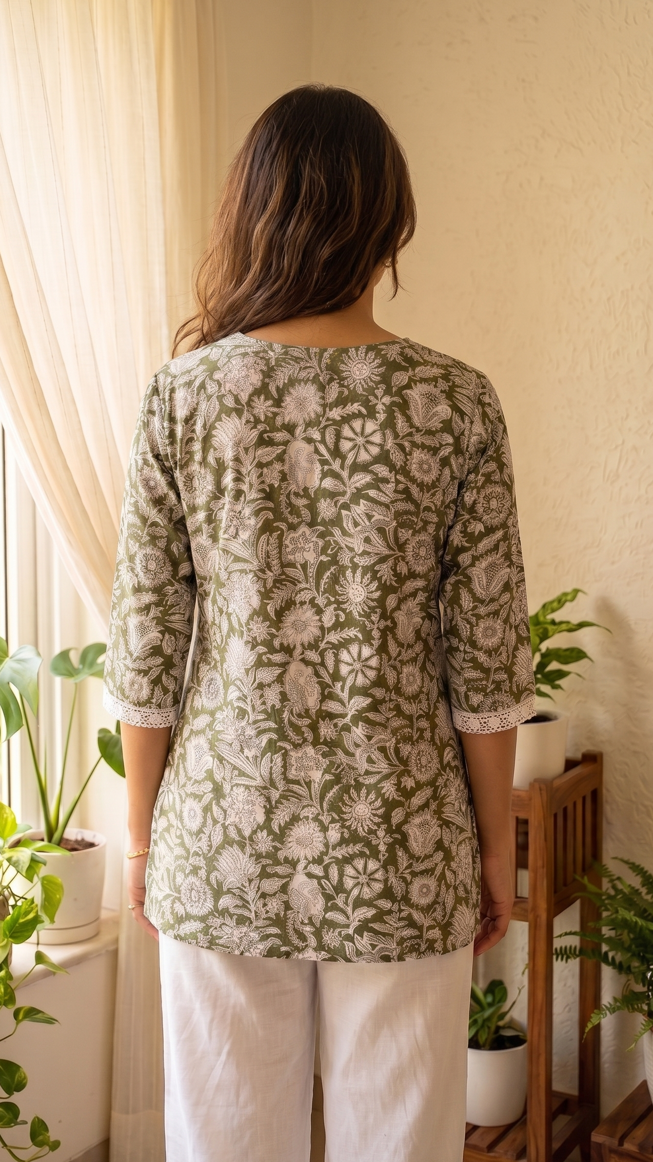 Woman wearing a green patterned top and white pants standing in a room with plants.