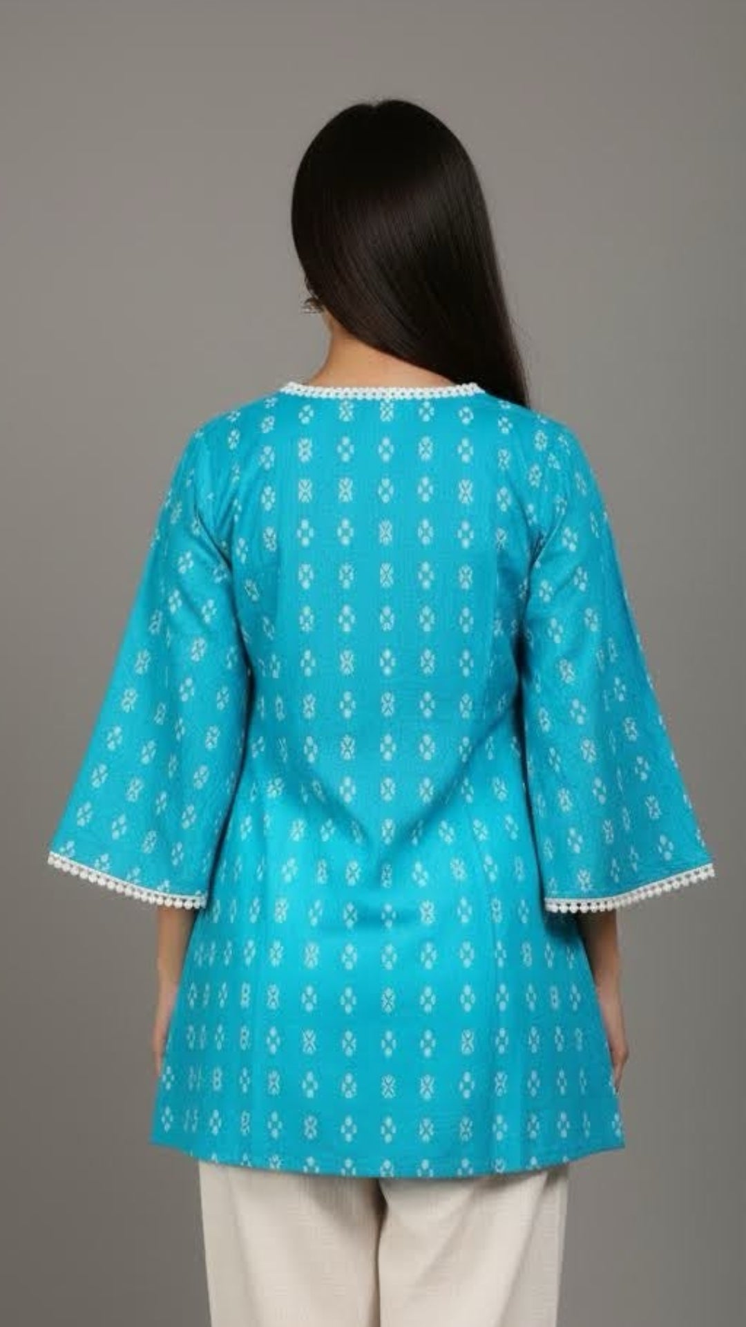 Woman wearing a turquoise top with white patterns and white pants on a gray background