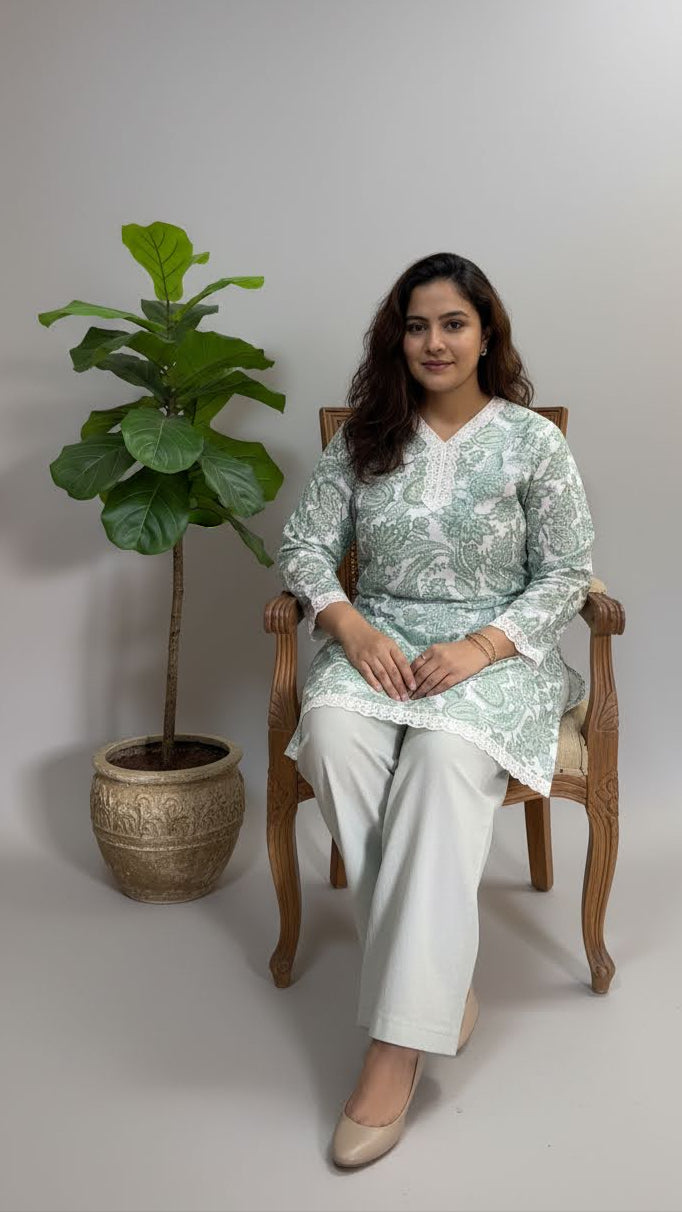 Woman sitting on a chair wearing a light green kurta with white pants, surrounded by a plant and decorative pot.