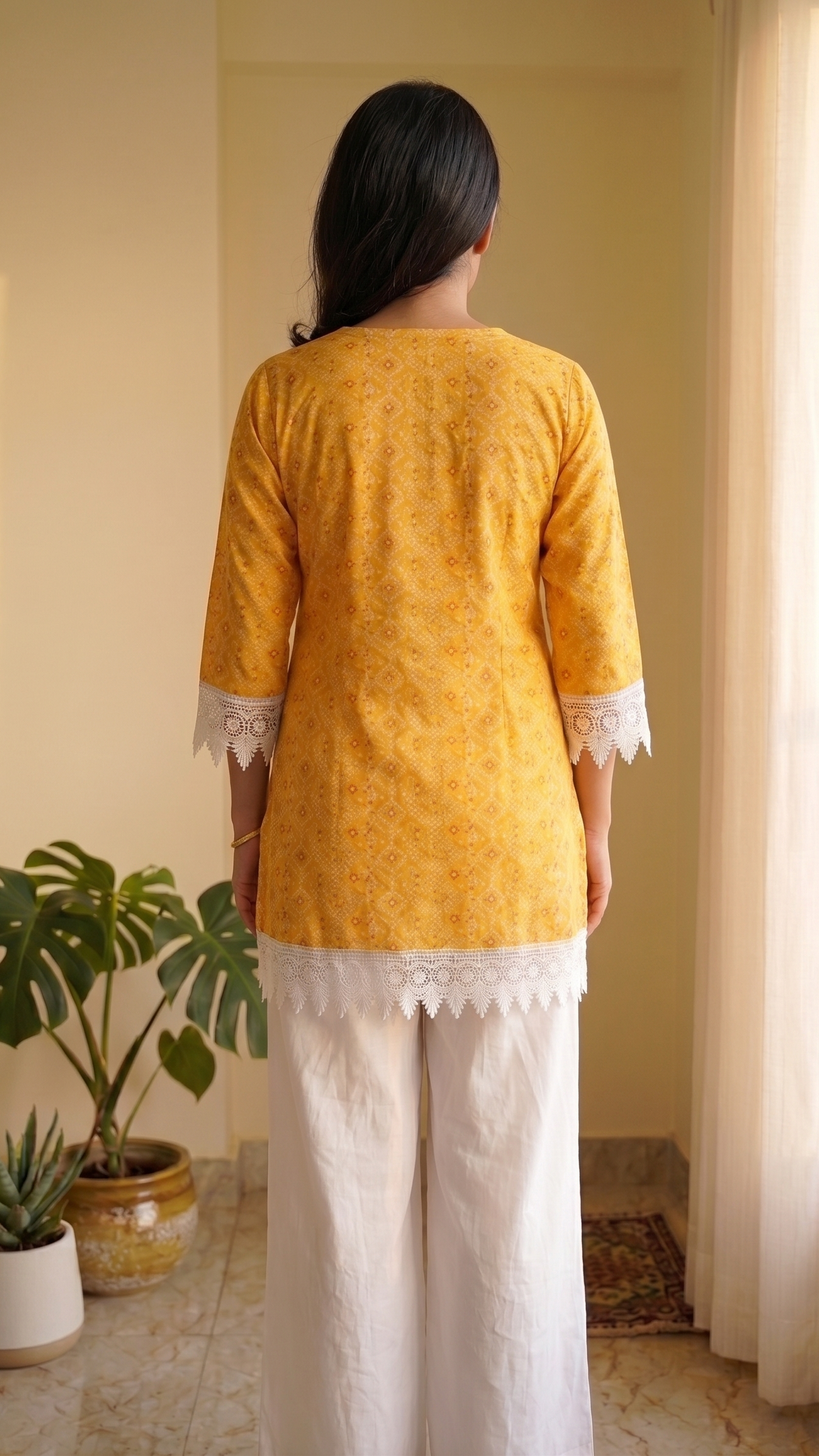 Person wearing a mustard yellow top and white pants standing in a room with plants and a window.