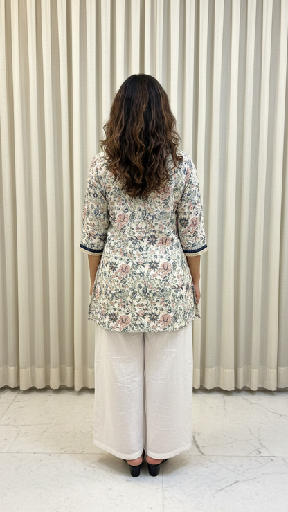 Woman wearing a floral blouse and white pants standing against a beige curtain.
