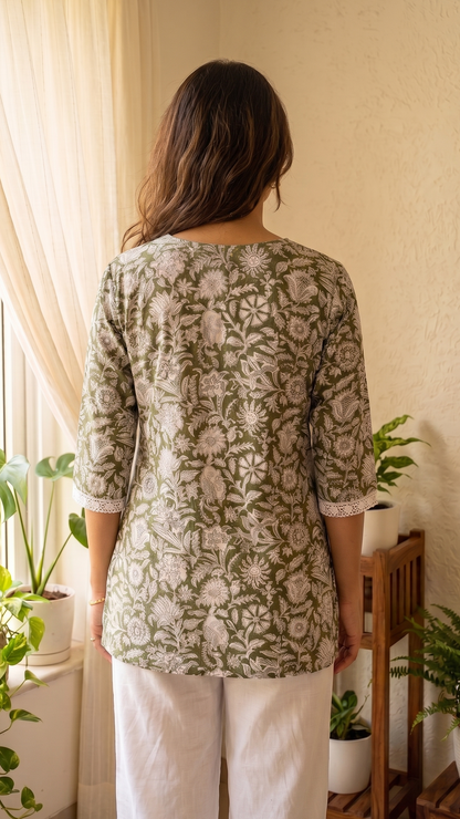 Woman wearing a green patterned top and white pants standing in a room with plants.