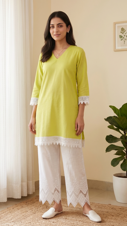 Woman wearing a green kurta with white lace details standing in a room with a plant and window.