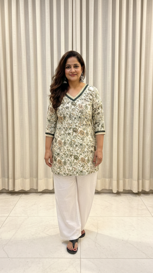 Woman wearing a floral top and white pants standing in front of a curtain.