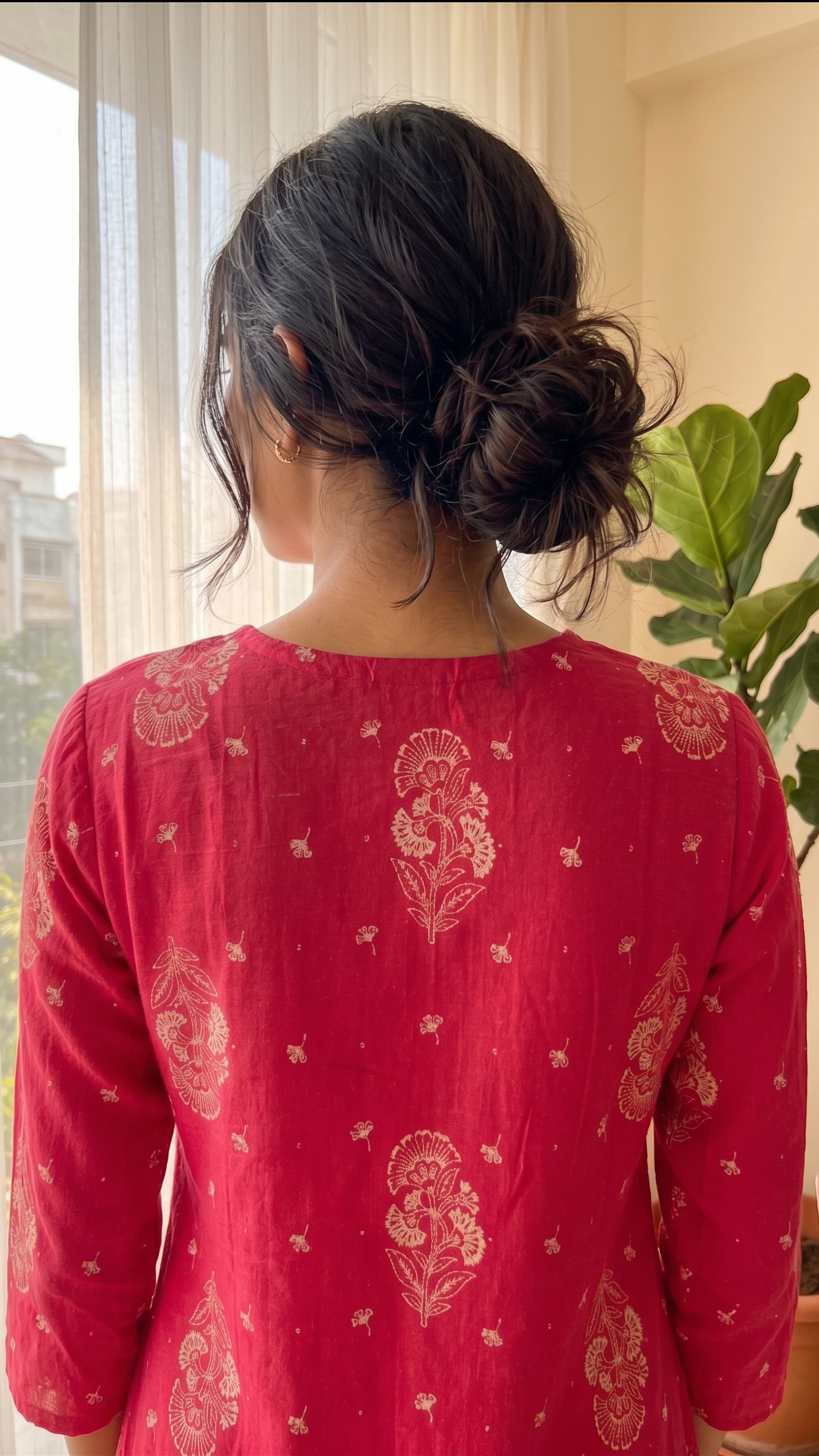 Person wearing a red patterned top standing by a window with a plant in the background