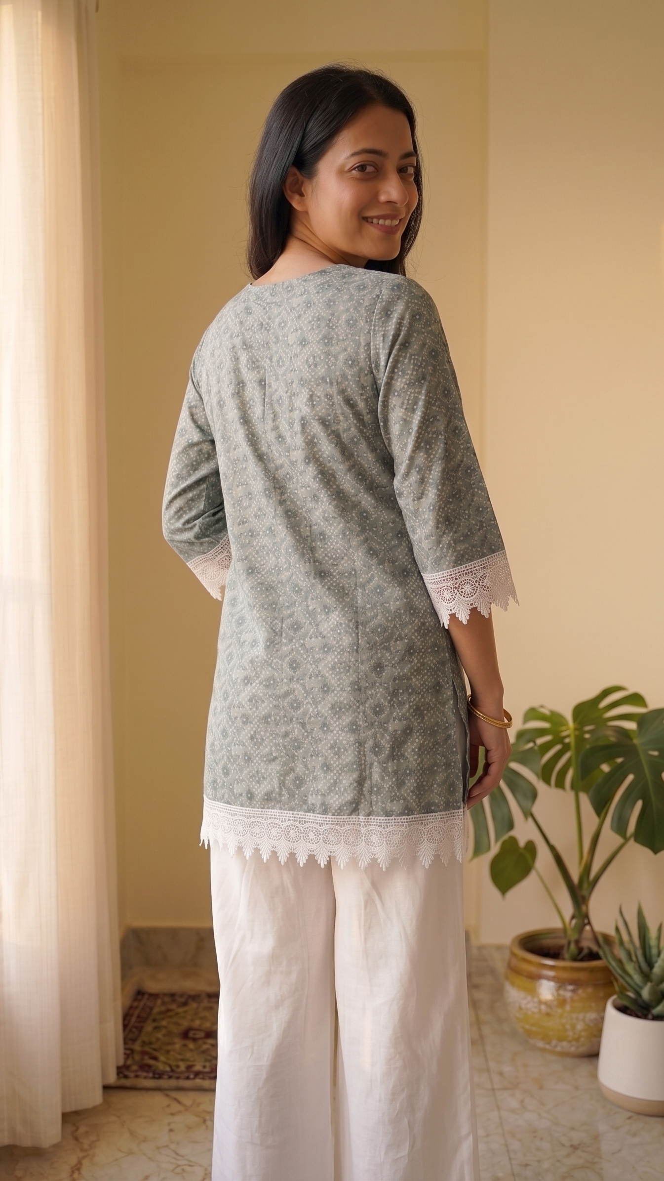 Woman wearing a green patterned top and white pants standing in a room with plants.