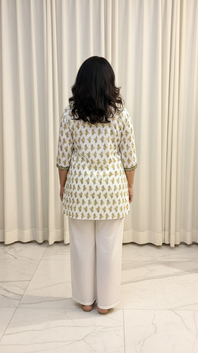 Woman wearing a patterned top and white pants standing in front of a white curtain.
