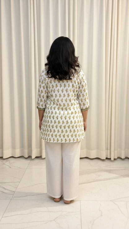 Woman wearing a patterned top and white pants standing in front of a white curtain.