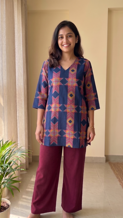Woman wearing a patterned top and maroon pants standing in a room with a plant and curtains.