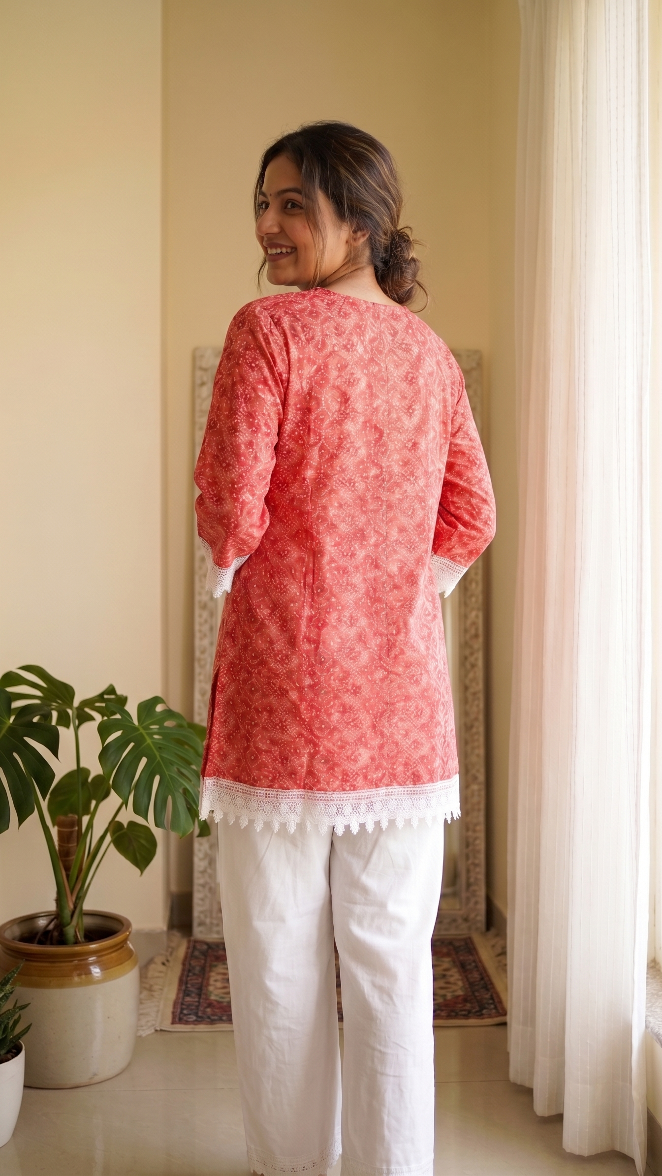 Woman wearing a red patterned top and white pants standing in a room with a plant and curtains.