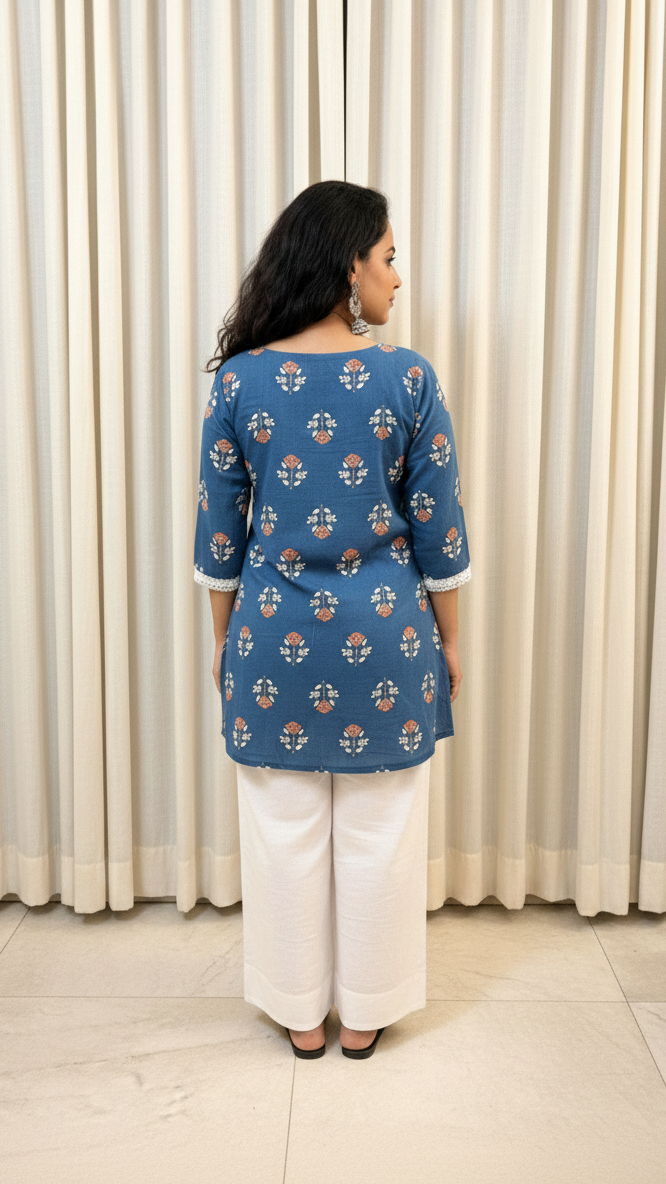 Woman wearing a blue patterned top and white pants standing against a white curtain.