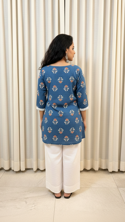 Woman wearing a blue patterned top and white pants standing against a white curtain.