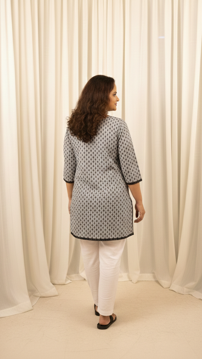 Woman wearing a patterned top and white pants standing in front of white curtains.