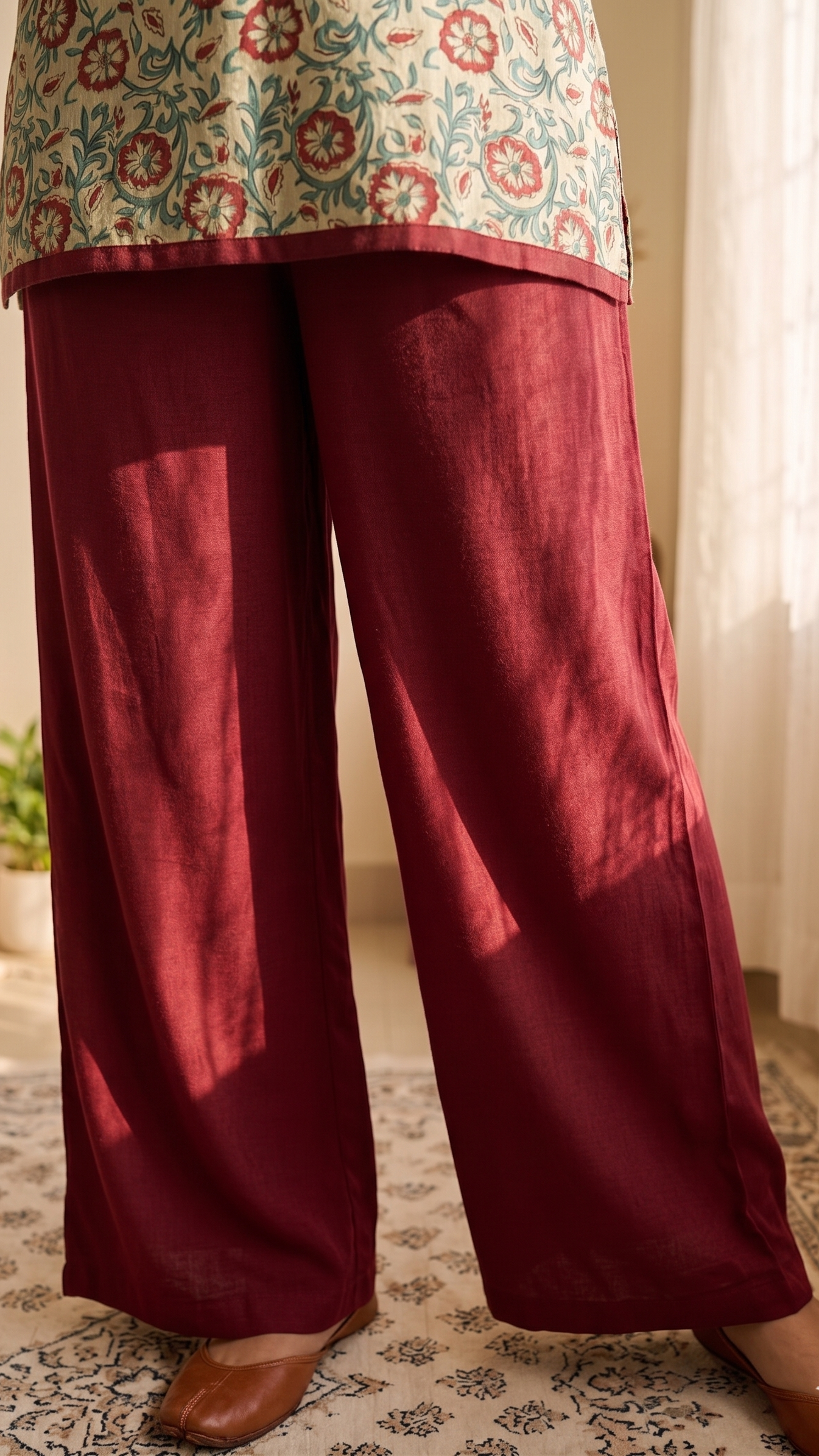 Maroon pants worn with a patterned top in a room with curtains and a plant.