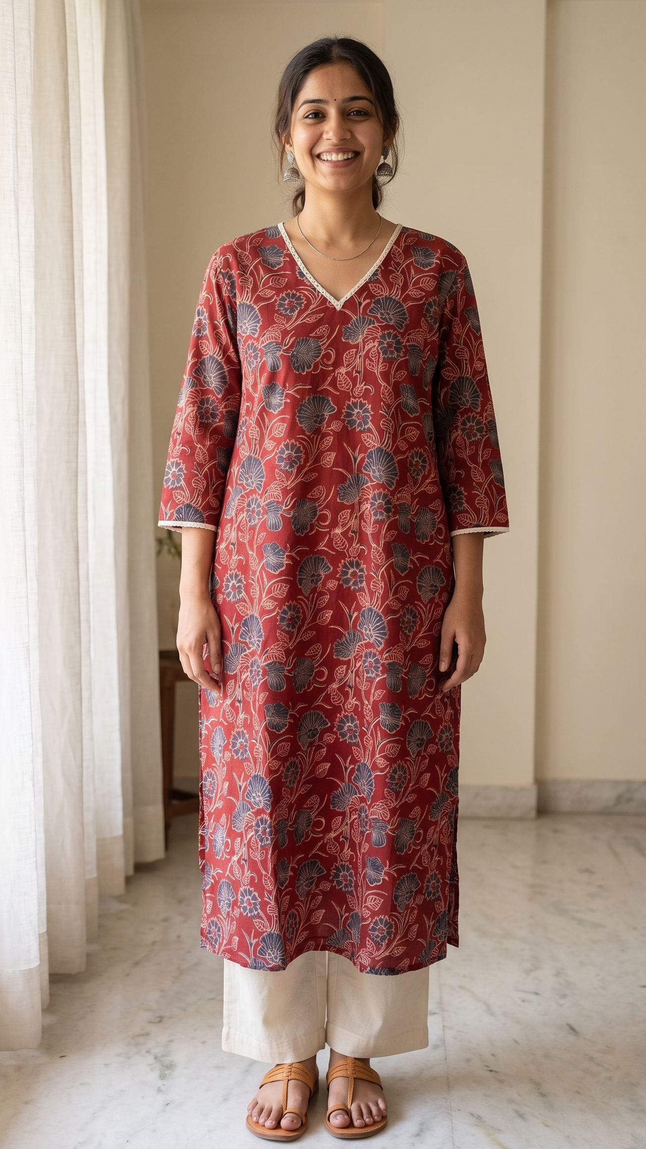 Woman wearing a red floral kurta standing in a room with light curtains and a plant.