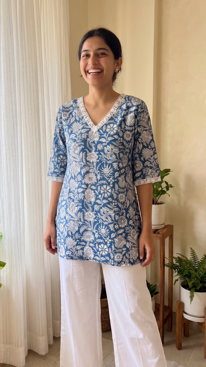 Woman wearing a blue floral top and white pants standing in a room with plants.
