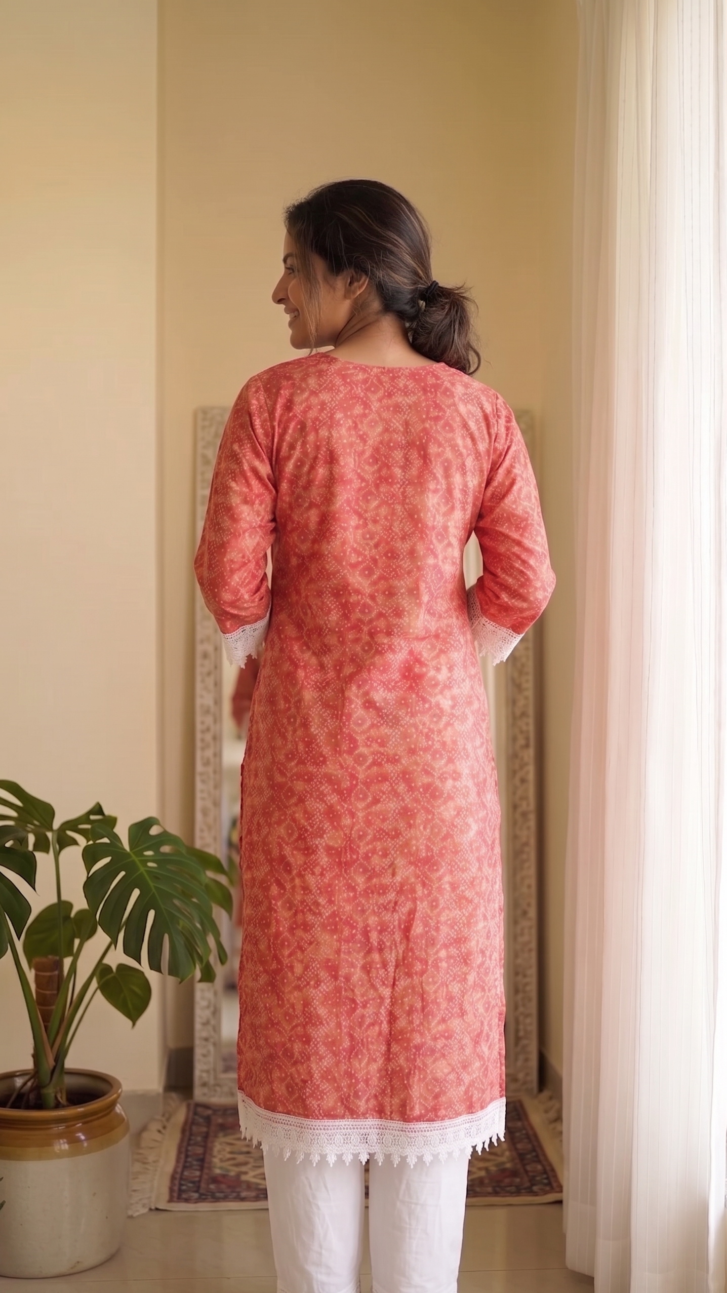 Woman wearing a pink kurta with white pants standing in a room with a plant and curtains.