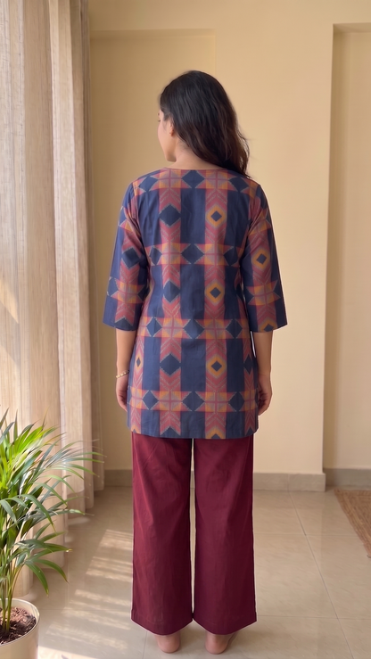 Woman wearing a patterned top and maroon pants standing in a room with a plant and window.