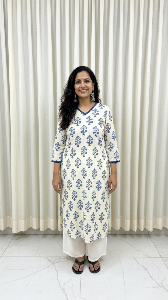 Woman wearing a white kurta with blue floral patterns standing against a white curtain.