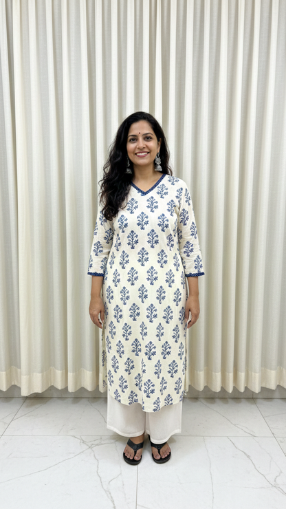Woman wearing a white kurta with blue floral patterns standing against a white curtain.
