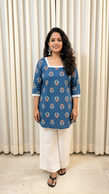 Woman wearing a blue floral top and white pants standing in front of beige curtains.