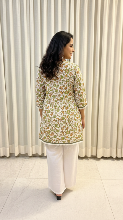 Woman wearing a floral top and white pants standing against a white curtain.