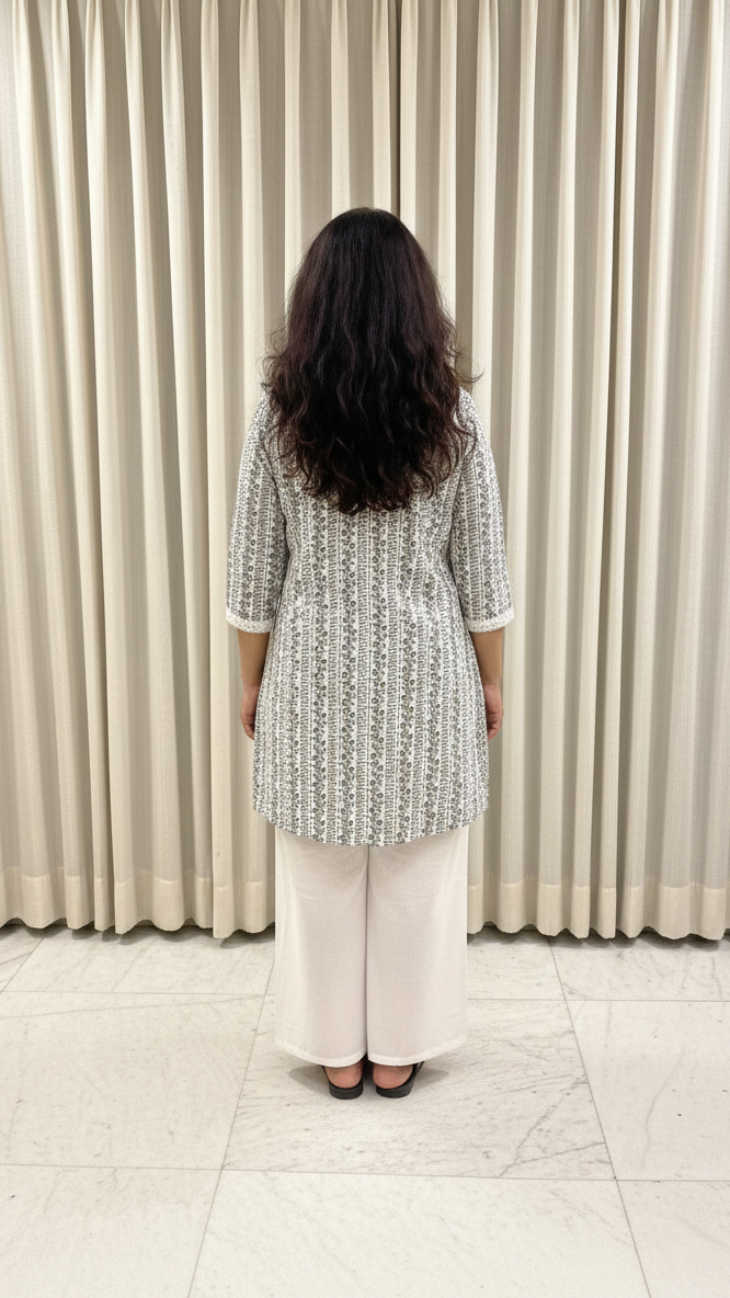 Woman wearing a patterned top and white pants standing in front of a beige curtain.
