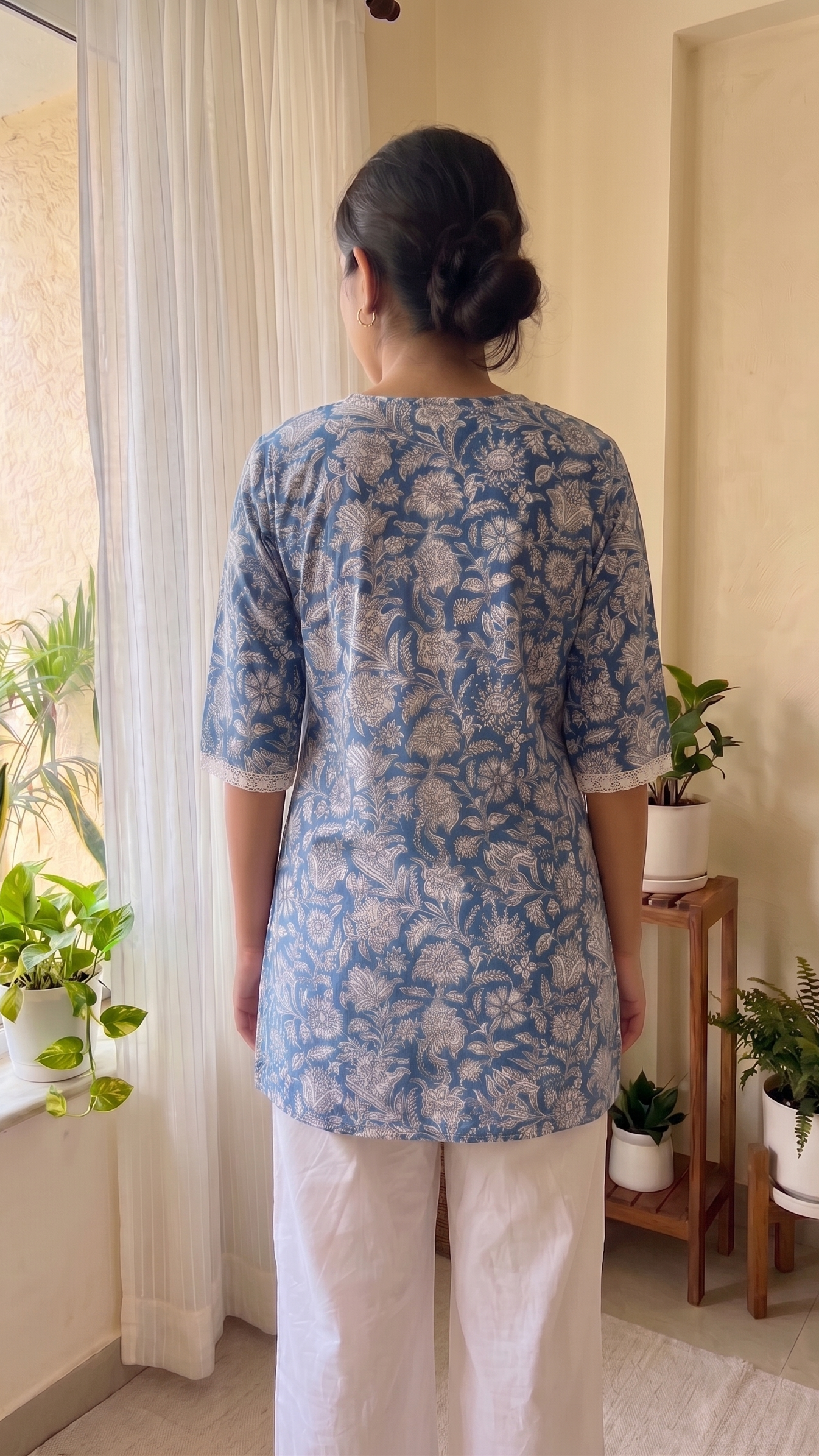 Person wearing a blue floral top and white pants standing in a room with plants.