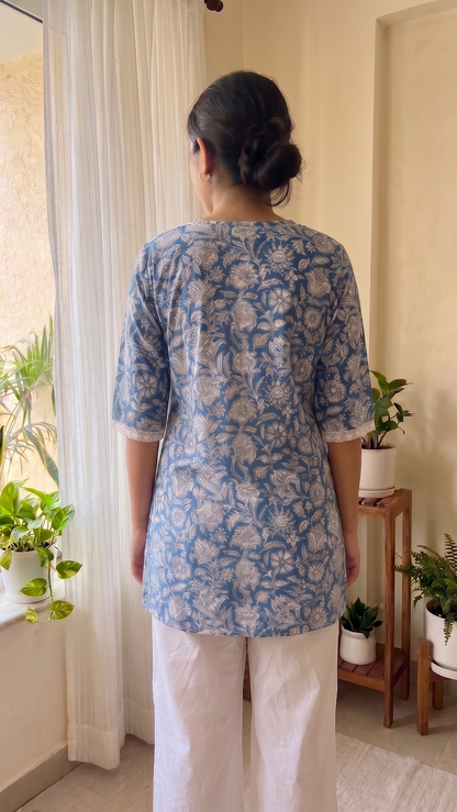 Person wearing a blue floral top and white pants standing in a room with plants.